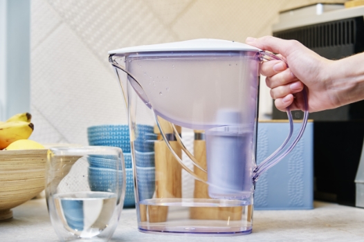 a person's hand holding a water filter pitcher with water in it next to a small glass of water